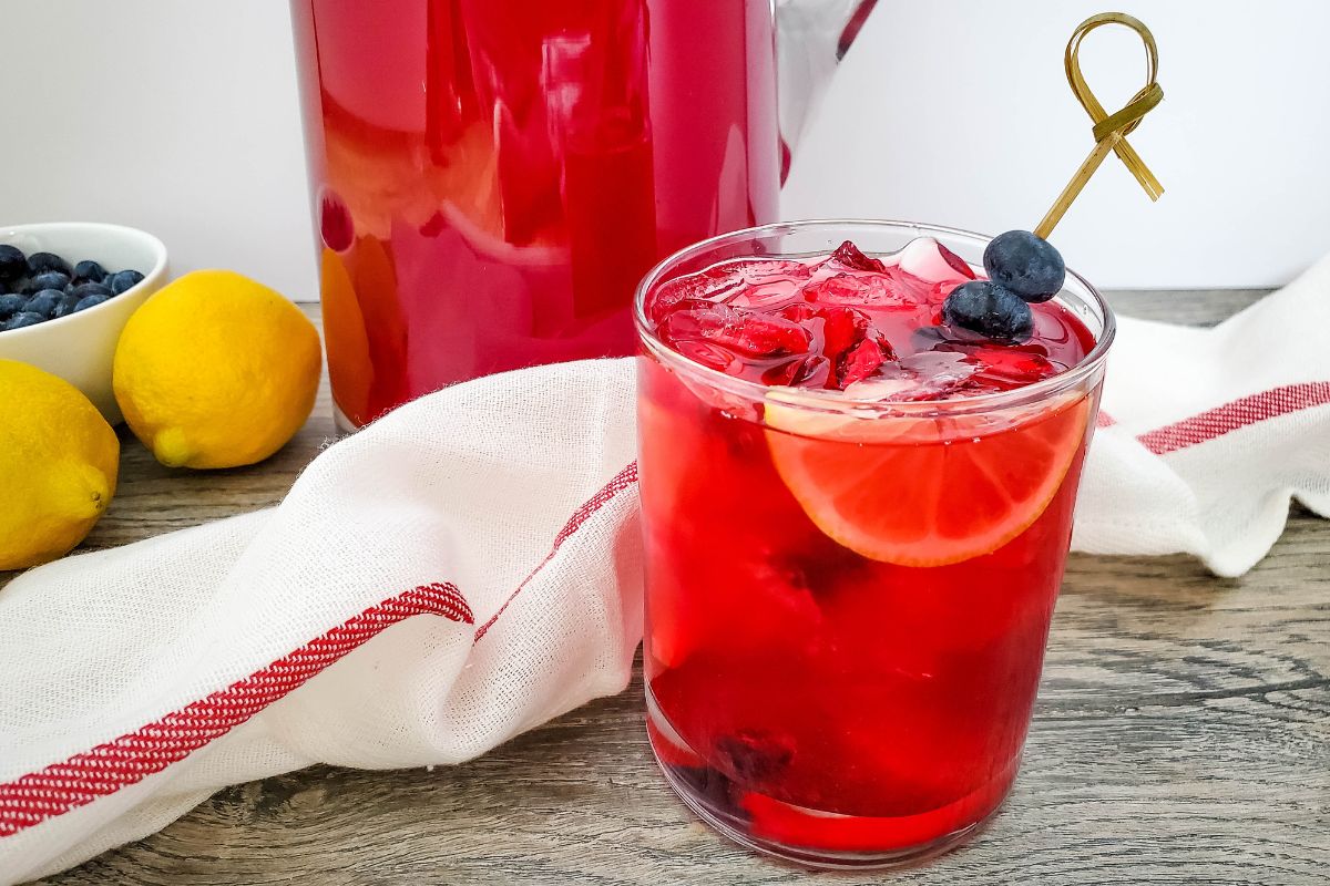 Blueberry Lemonade served in a glass with a wedge of lemon and a skewer of blueberries. In the background is the pitcher of lemonade and a bowl of fresh lemons and blueberries