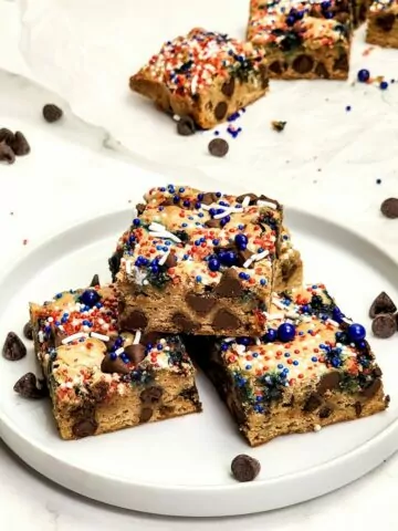 Chocolate chip cookie bars with red white and blue sprinkles on top stacked on a white plate.