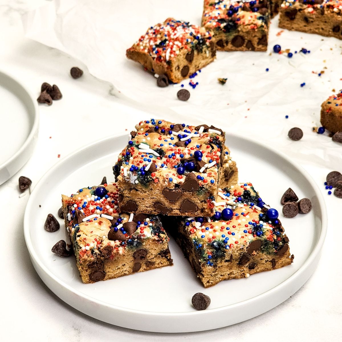 Chocolate chip cookie bars with red white and blue sprinkles on top stacked on a white plate.