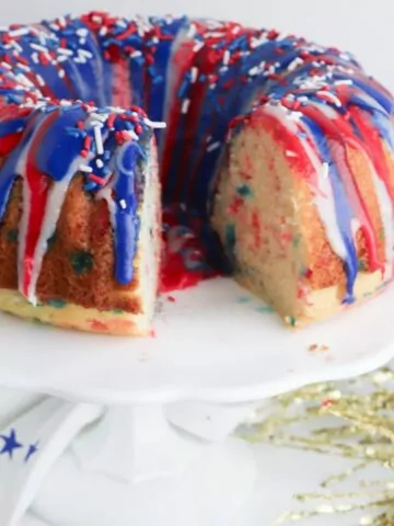 Red white and blue funfetti bundt cake and frosting with patriotic sprinkles on top. The pound cake is displayed on a white cake stand with a slice removed to show the inside of the cake.