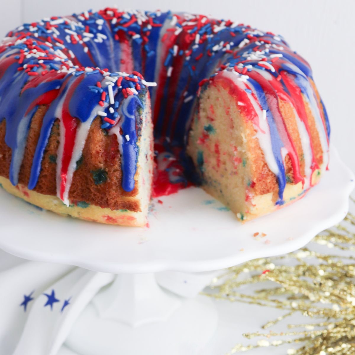 Red white and blue funfetti bundt cake and frosting with patriotic sprinkles on top. The pound cake is displayed on a white cake stand with a slice removed to show the inside of the cake.