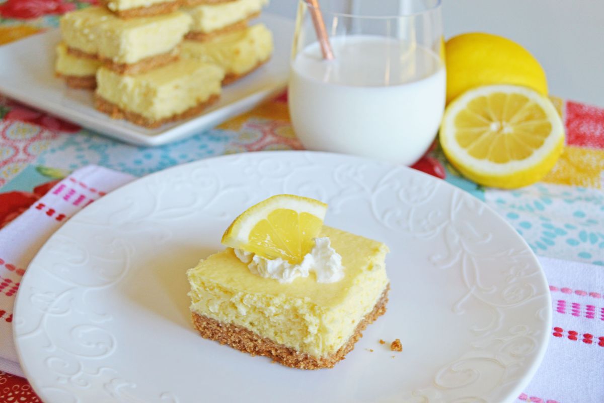 lemon cheesecake bar topped with whipped cream and lemon wedge. Glass of milk and cheesecake bars in the background