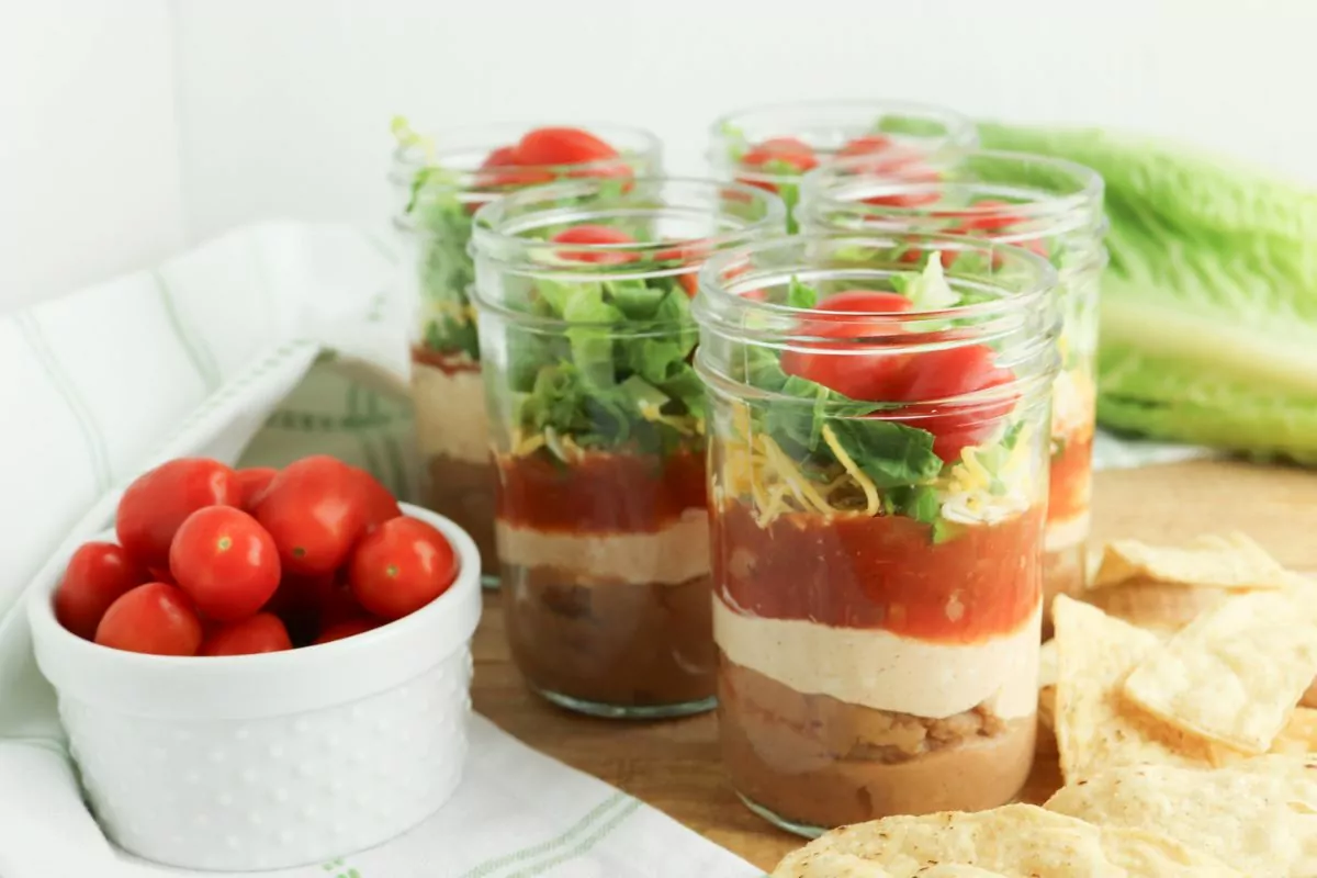 Mason Jar Taco Dip with a bowl of cherry tomatoes, chips, and romaine around the jars.
