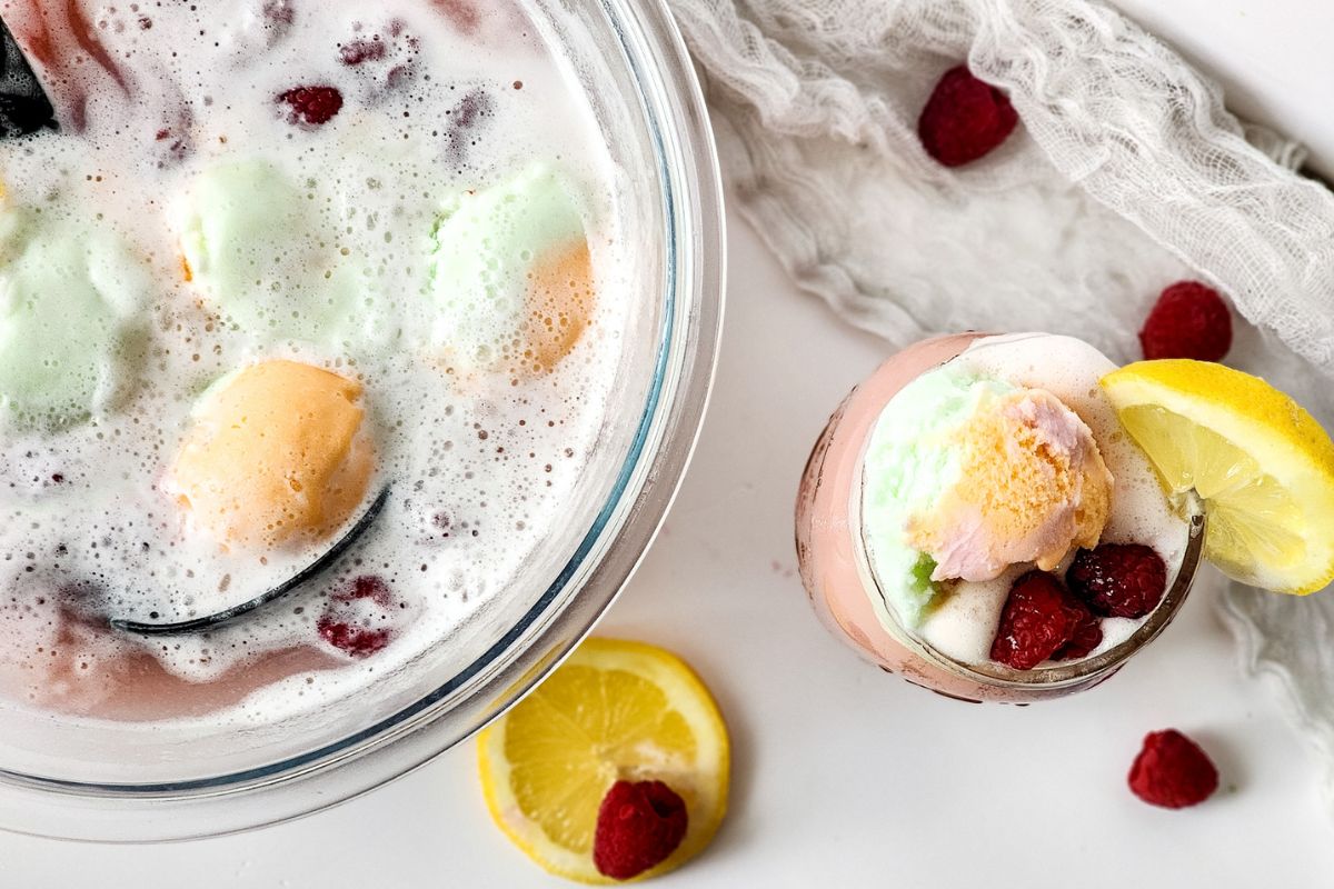 punch bowl filled with raspberry lemonade sherbet punch with a filled cup garnished with fresh lemon and raspberries