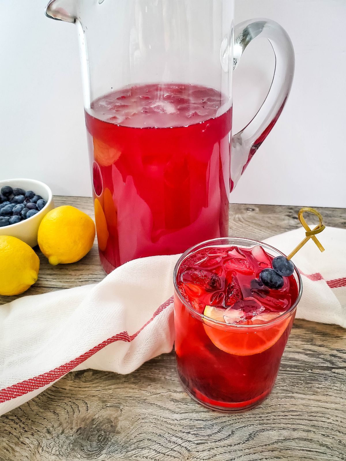 Blueberry Lemonade served in a glass with a wedge of lemon and a skewer of blueberries. The pitcher of lemonade and fresh blueberries and lemons are in the background