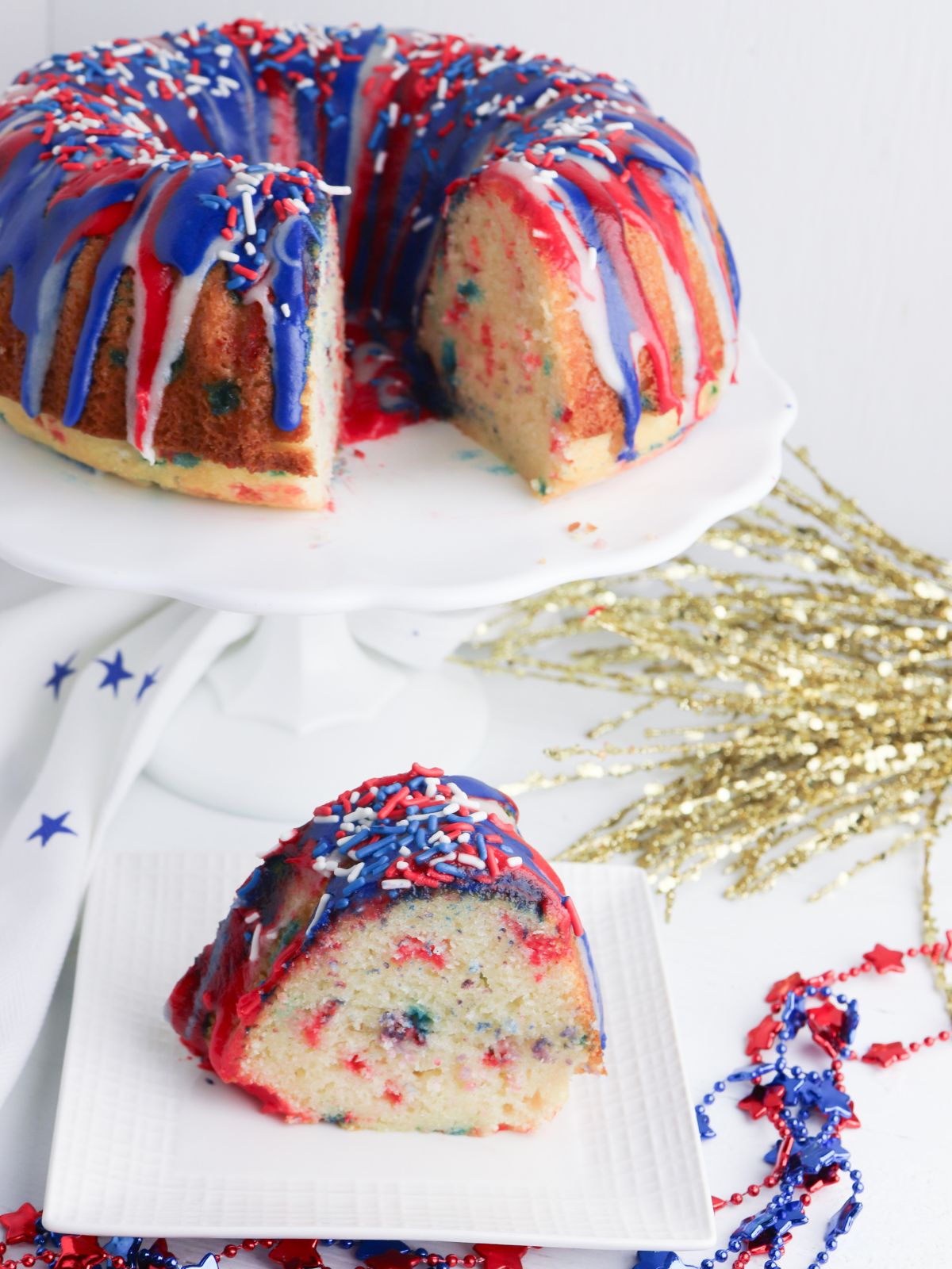 Slice of red and blue confetti cake on a square plate with the rest of the cake on a cake stand in the background.