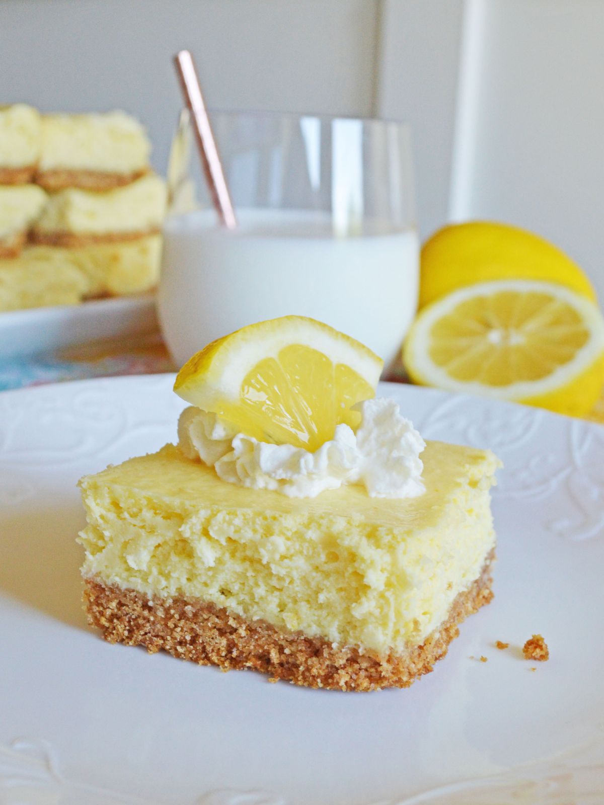 lemon cheesecake bars with graham cracker crust. Background has a plate of the bars, a glass of milk, and fresh lemons