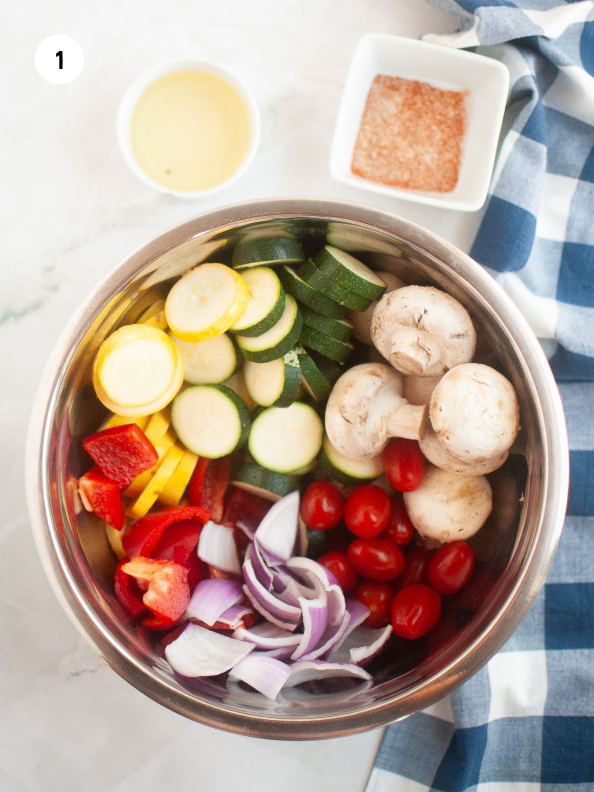 mixing bowl filled with all the cut up vegetables. Above the bowl are small bowls of olive oil and seasonings.