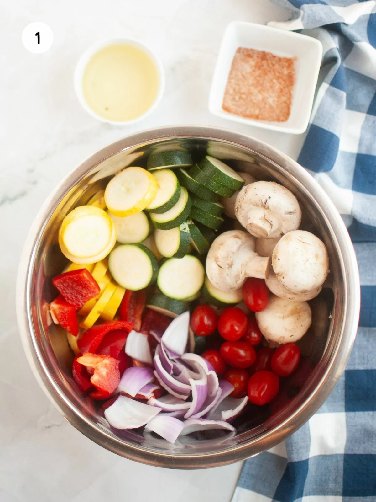 mixing bowl filled with all the cut up vegetables. Above the bowl are small bowls of olive oil and seasonings.