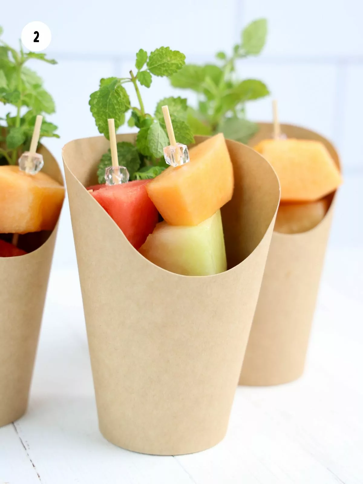 Fruit skewers and lemon balm sprigs in brown charcuterie cups.