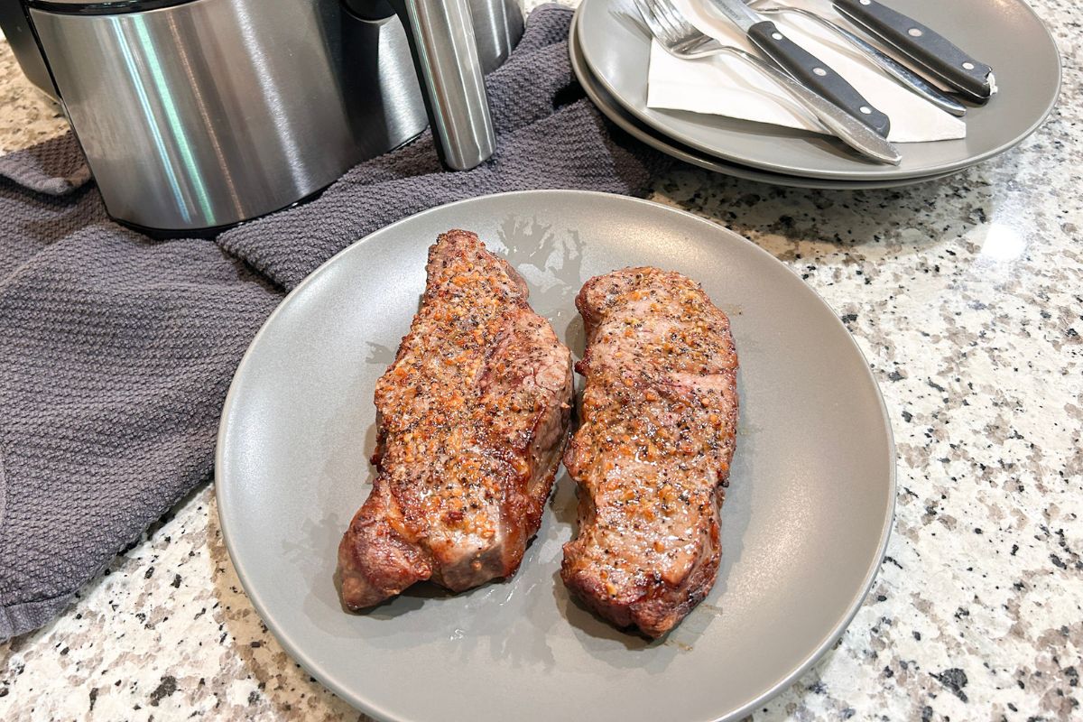 Two New York Strip steaks served on a gray plate. The air fryer and plates with silverware are in the background.