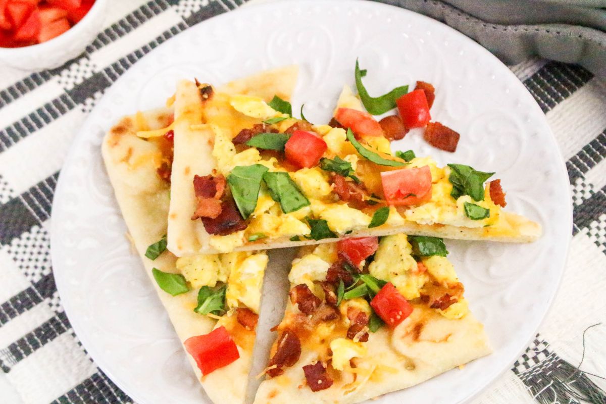 Slices of flatbread with scrambled eggs, bacon, cheese, and fresh tomatoes and spinach on top.