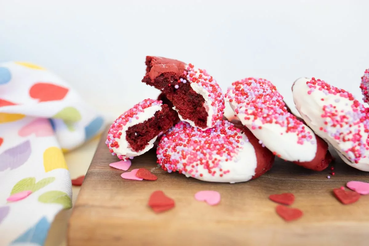 Red velvet cookies dipped in white chocolate on a wood board. One cookie is broken in half to show the inside.