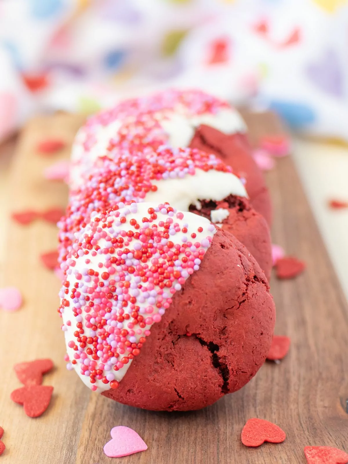 Red velvet cookies dipped in white chocolate and pink sprinkles. Red heart sprinkles are scattered around the cookies.