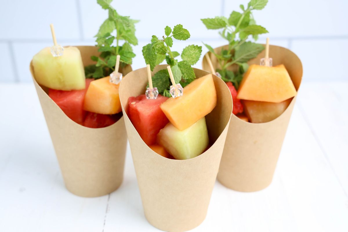 3 brown charcuterie cups filled with skewers of watermelon, cantaloupe, and honeydew melon and a sprig of lemon balm.