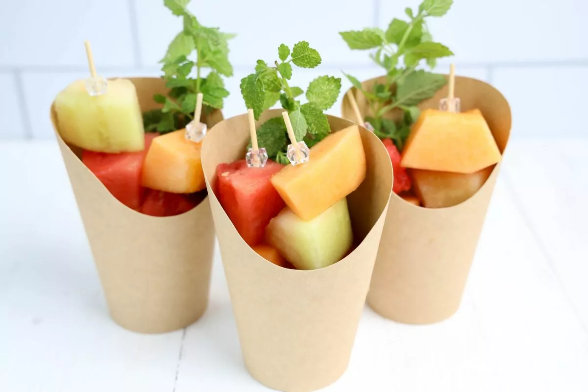 3 brown charcuterie cups filled with skewers of watermelon, cantaloupe, and honeydew melon and a sprig of lemon balm.