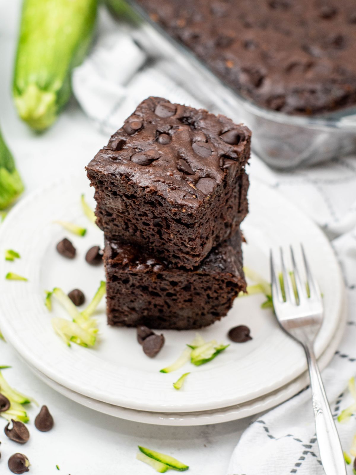Two slices of chocolate zucchini cake stacked on a white plate. A fresh zucchini and the rest of the cake is in the background.