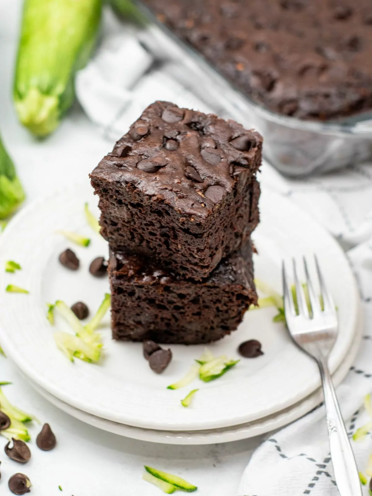 Two slices of chocolate zucchini cake stacked on a white plate. A fresh zucchini and the rest of the cake is in the background.