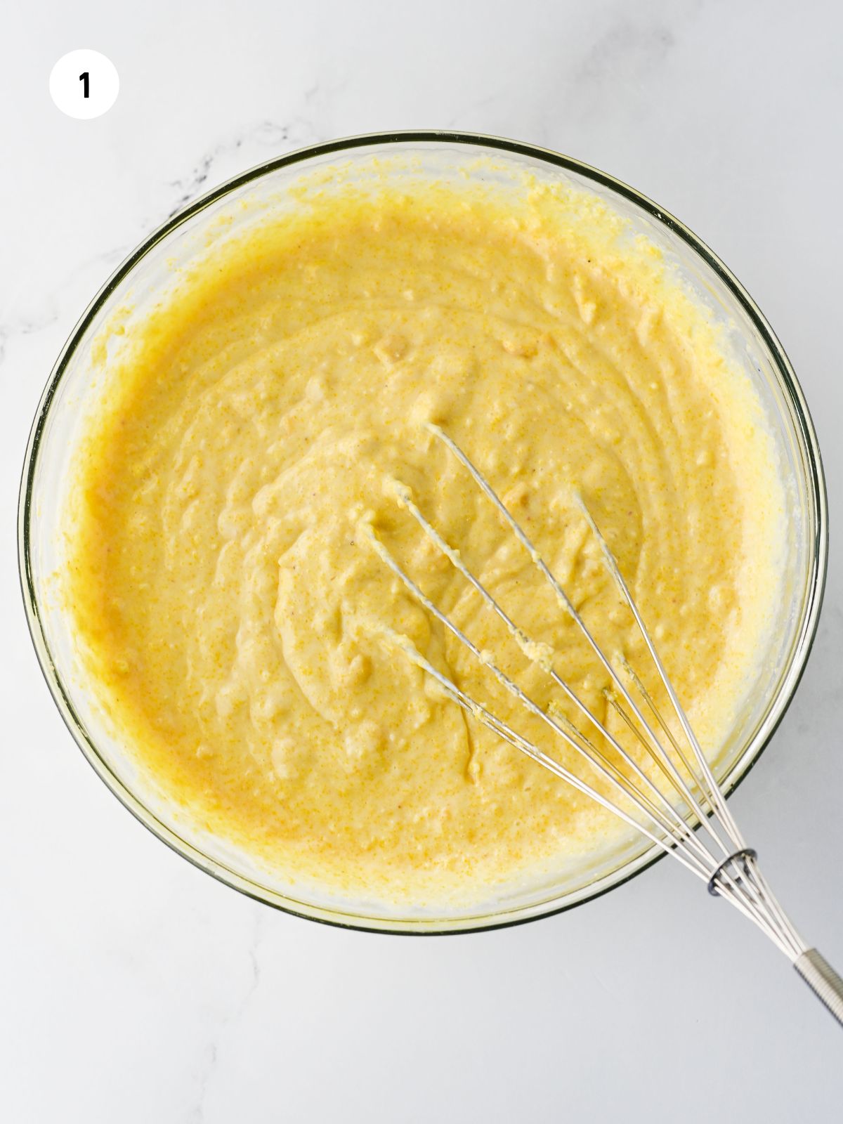 Cornbread batter in a clear mixing bowl. Whisk is in the batter.