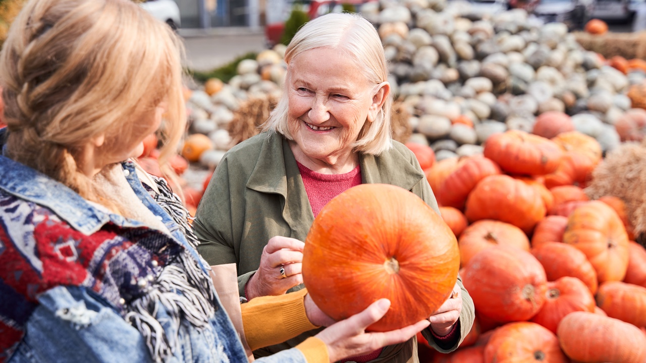 Blonde woman holding ripe orange pumpkin and showing it to her mother while choosing pumpkin at the autumn market