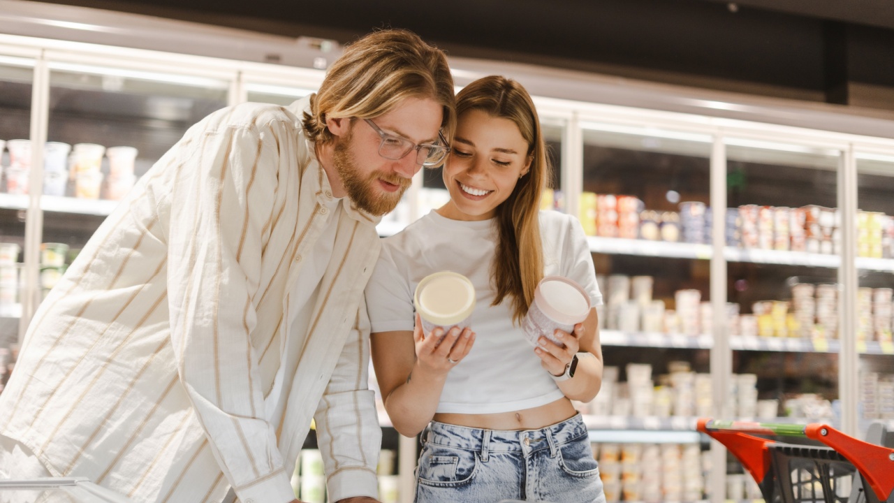 Happy young couple shopping for groceries, reading a product label while choosing frozen food from a supermarket fridge, doing grocery shopping together
