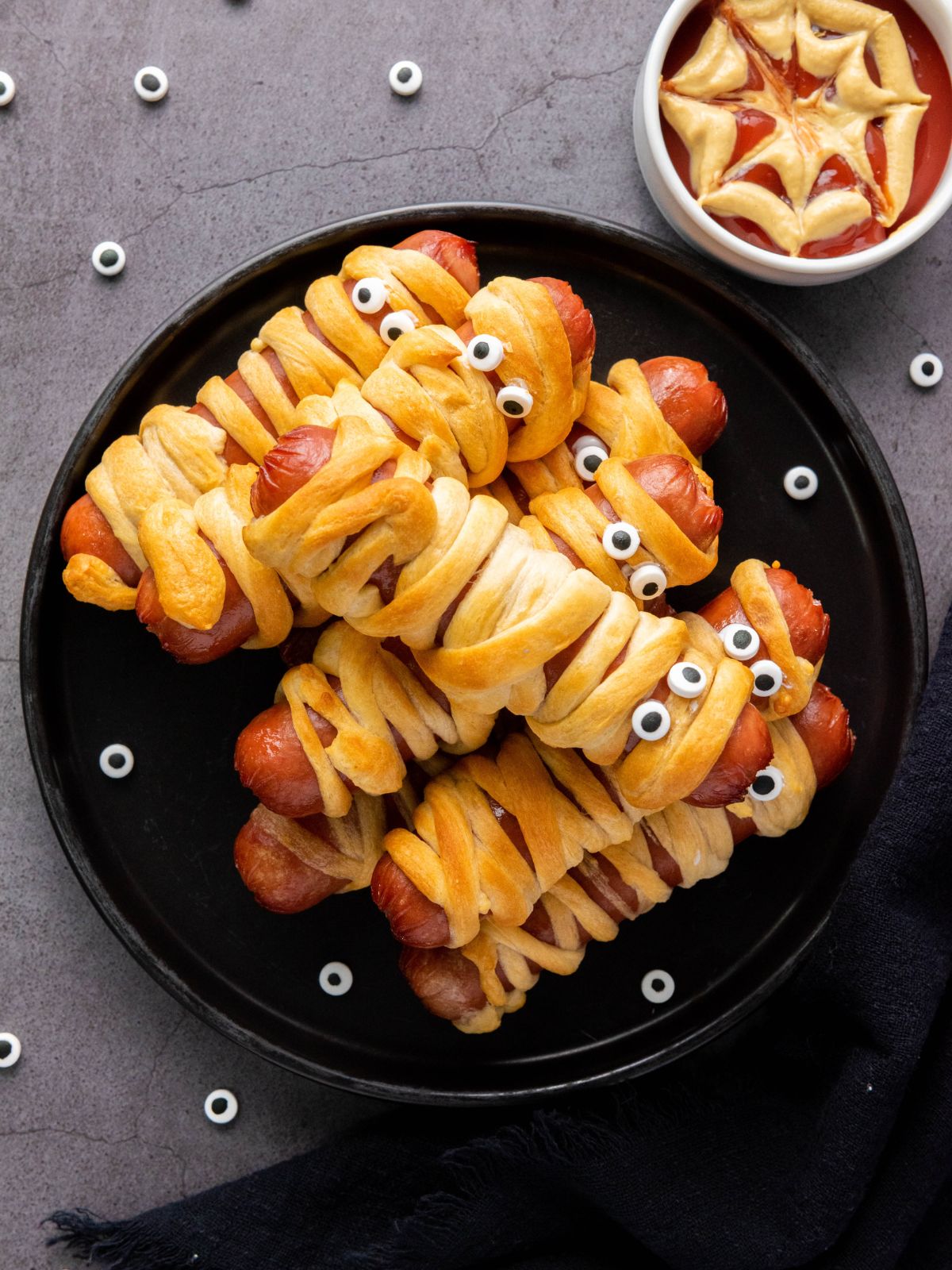 Plate of mummy hot dogs with crescent rolls and a bowl of ketchup and mustard in a spiderweb design.
