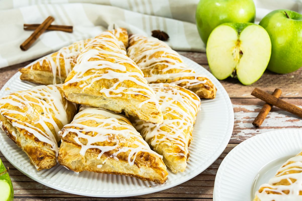 Plate of apple turnovers with glaze drizzled on top. Granny Smith apples in the background.