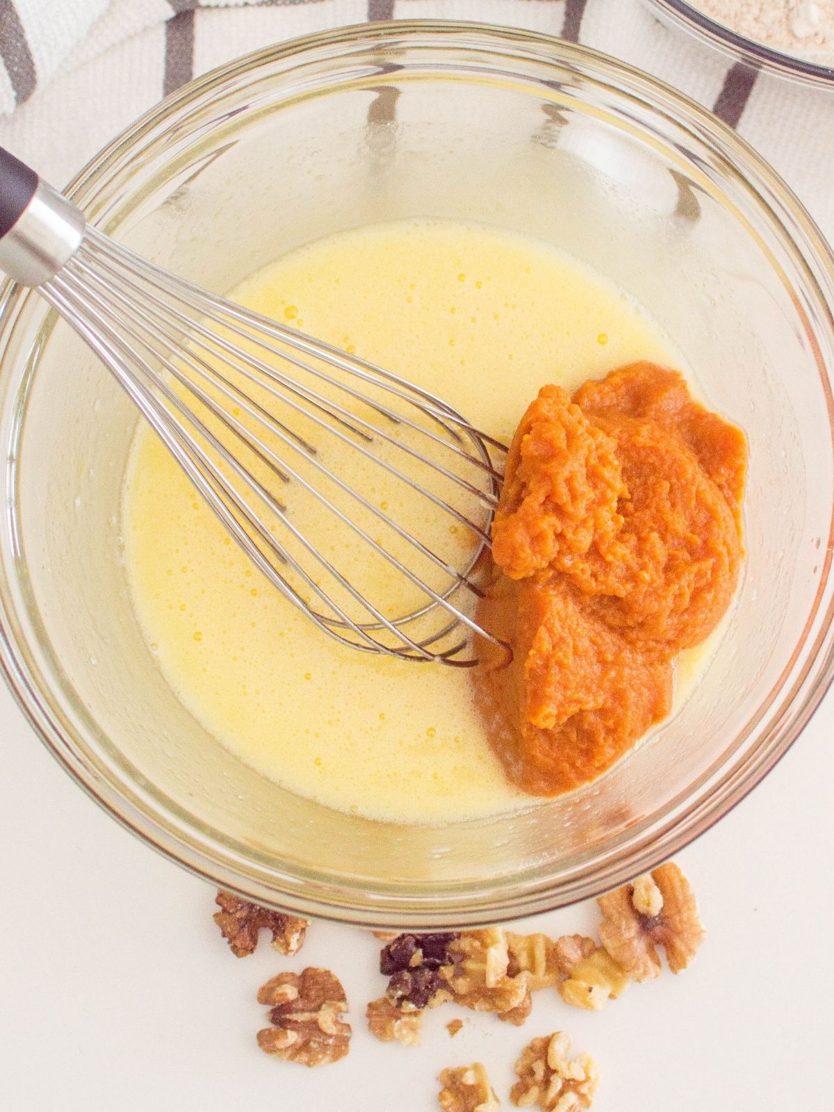 egg mixture with pumpkin puree in bowl with whisk. Walnuts on the tabletop in front of the bowl.