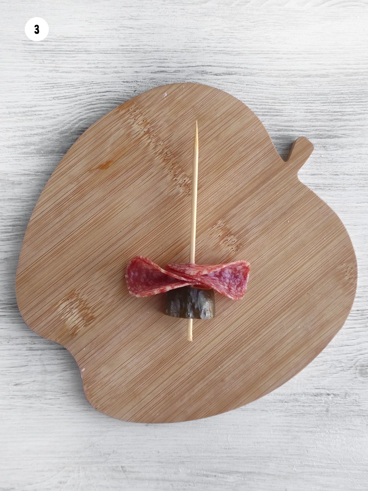 Skewer with a slice of pickle and salami ribbon on an apple shaped wood cutting board.