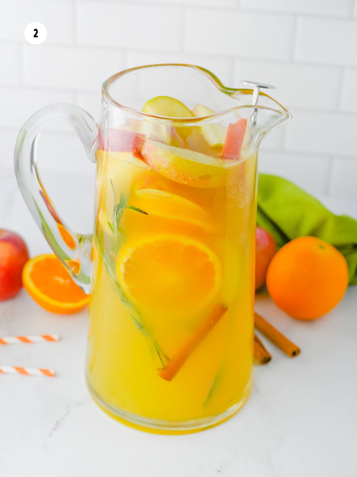pitcher of non-alcoholic beverage with oranges, apples, cinnamon sticks and sprigs of rosemary.