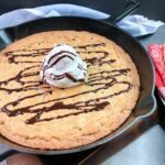 baked chocolate chip cookie in a cast iron skillet with scoop of whipped topping and chocolate sauce.