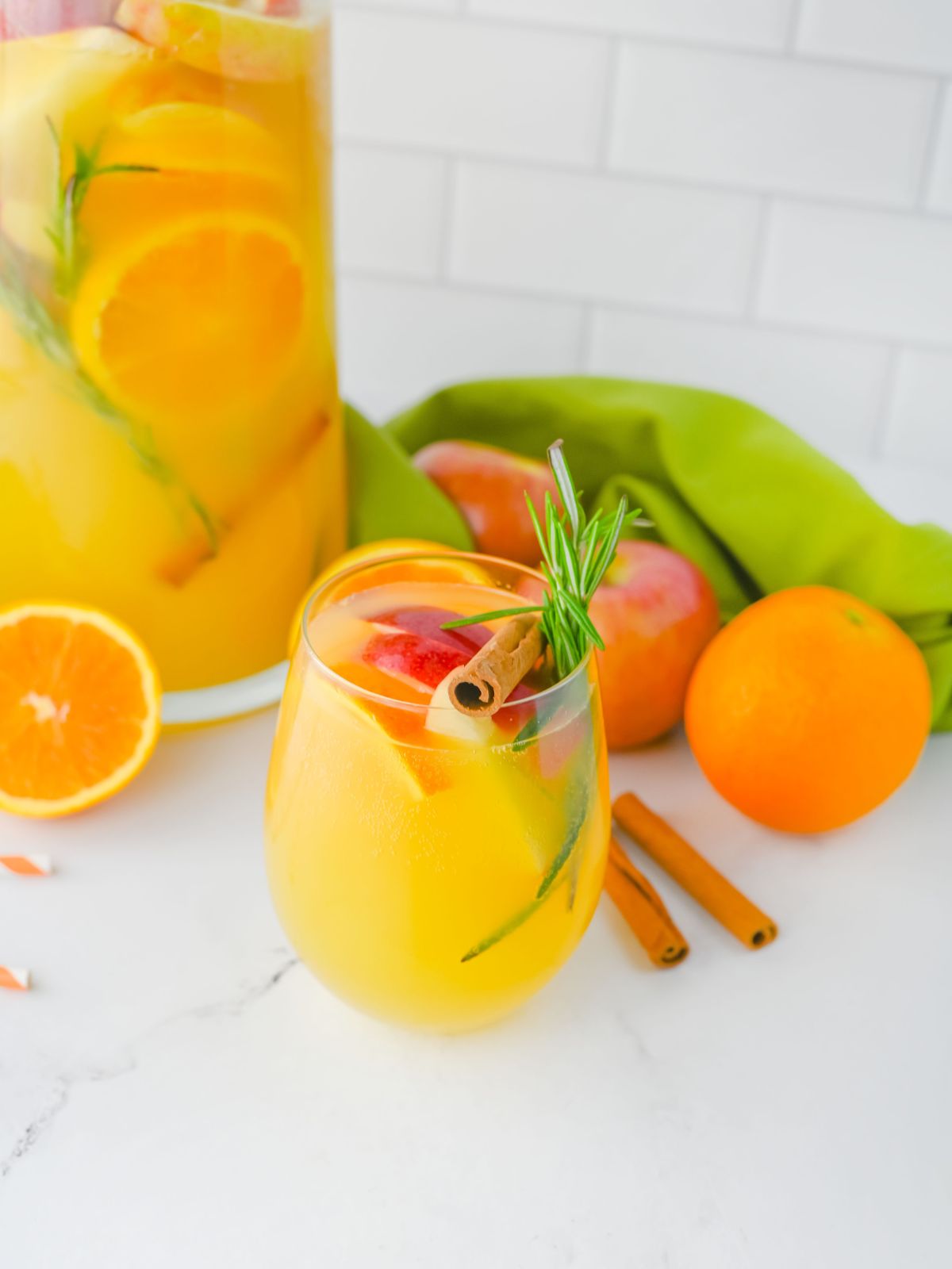 glass of non alcoholic beverage with orange juice, apples, oranges, cinnamon sticks and rosemary. Pitcher of punch in background.