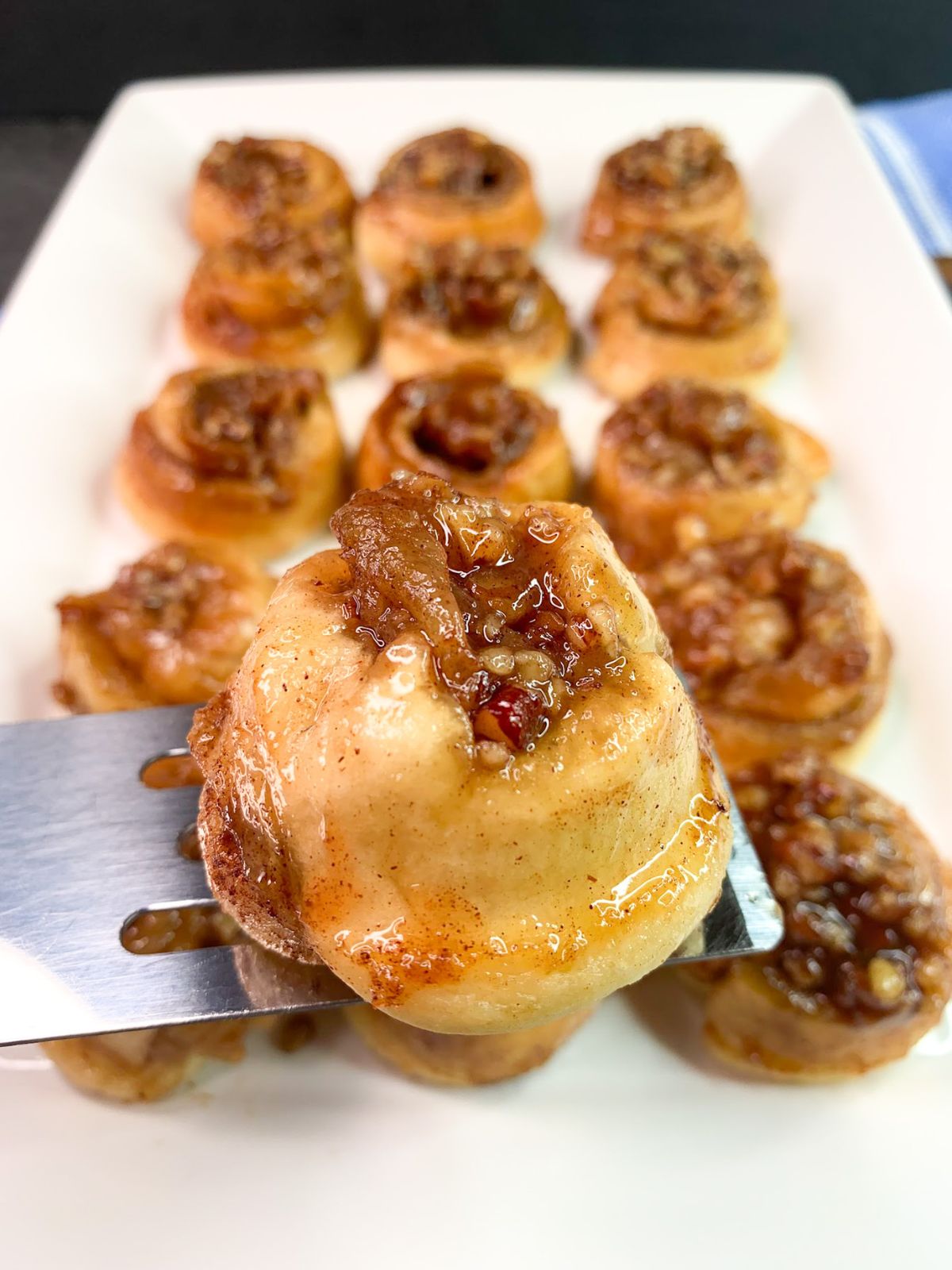 single serving pecan sticky bun on small spatula over a plate full of rolls.