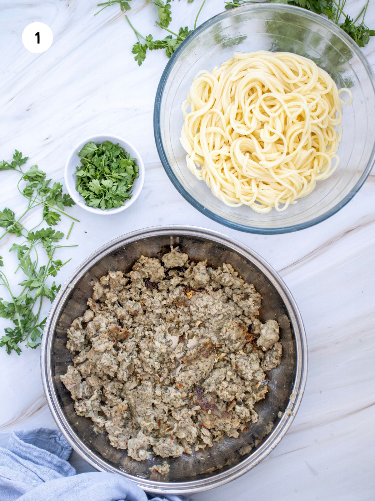 Pan of cooked Italian sausage, clear bowl with cooked spaghetti, and small bowl of chopped basil.