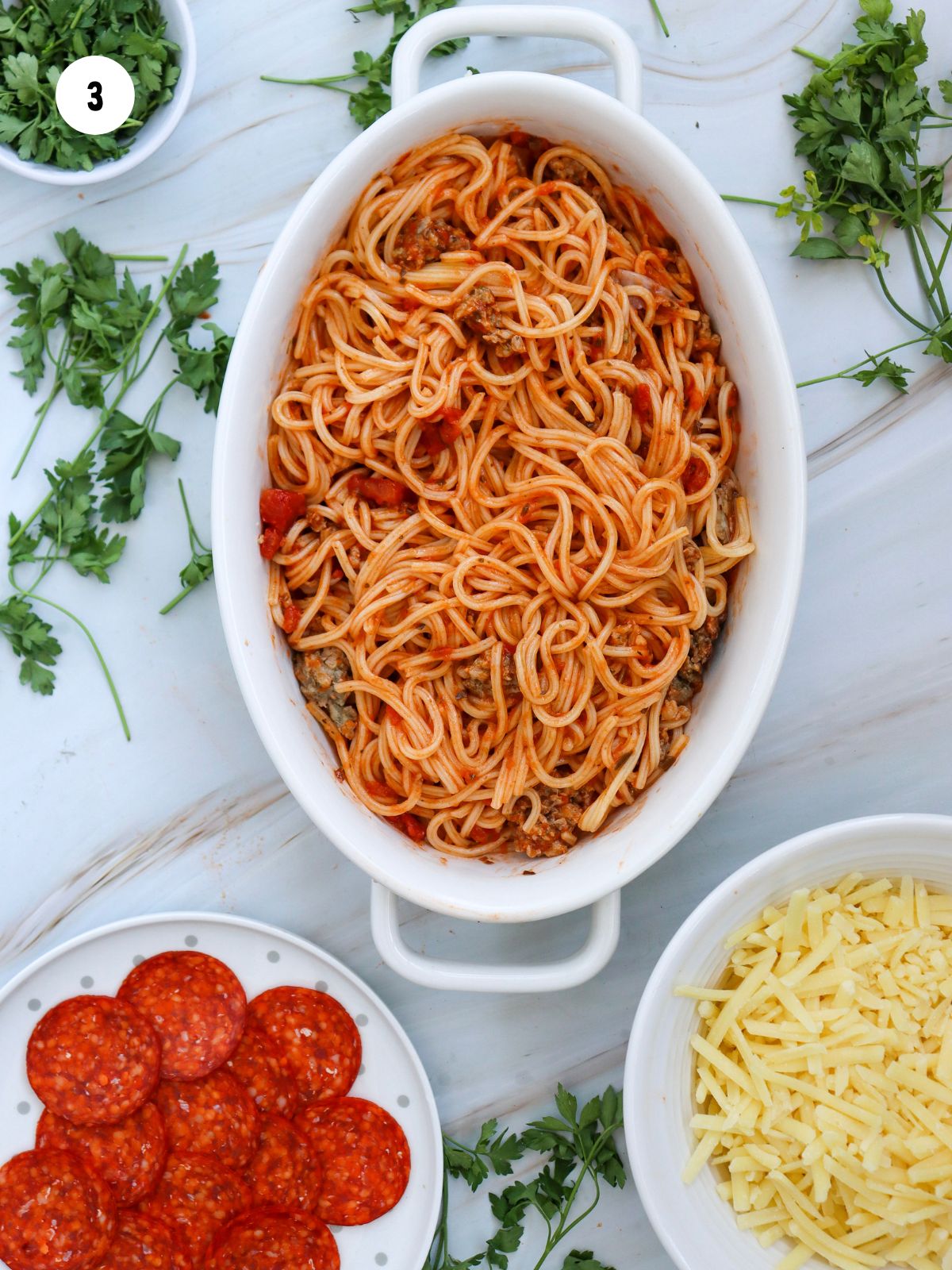 Oval casserole dish filled with spaghetti and meat mixed with the sauce. Plate of pepperoni and bowl of mozzarella.