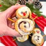 Closeup of Snowmen shaped cinnamon rolls with pretzel stick arms, chocolate chip eyes and a candy red nose.