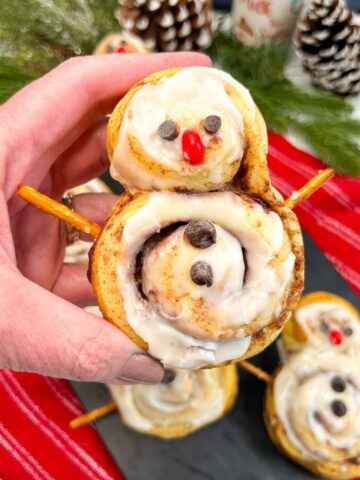Closeup of Snowmen shaped cinnamon rolls with pretzel stick arms, chocolate chip eyes and a candy red nose.