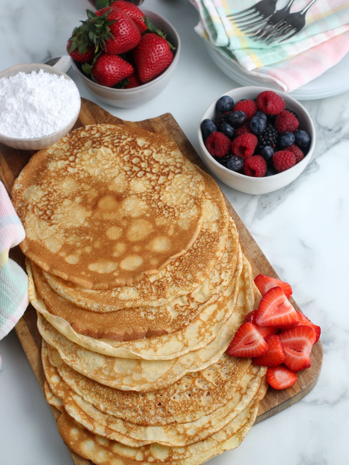 Homemade crepes on a serving board with bowls of fresh fruit and powdered sugar.