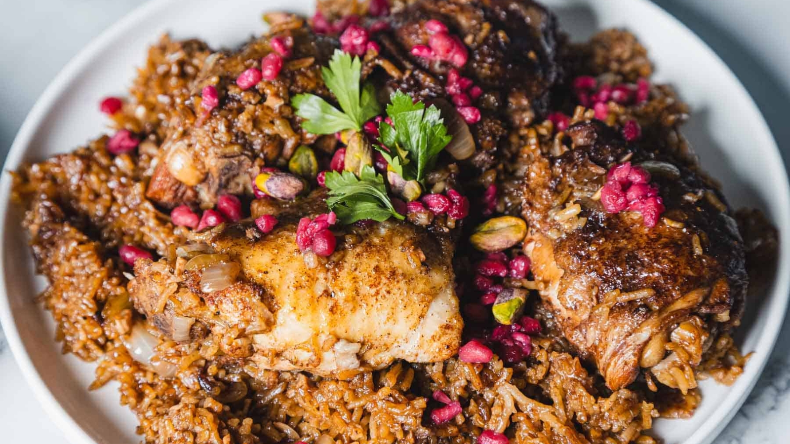 A close-up of a dish featuring seasoned rice topped with cooked chicken pieces. The meal is garnished with fresh parsley, pomegranate seeds, and pistachios, served on a white plate.