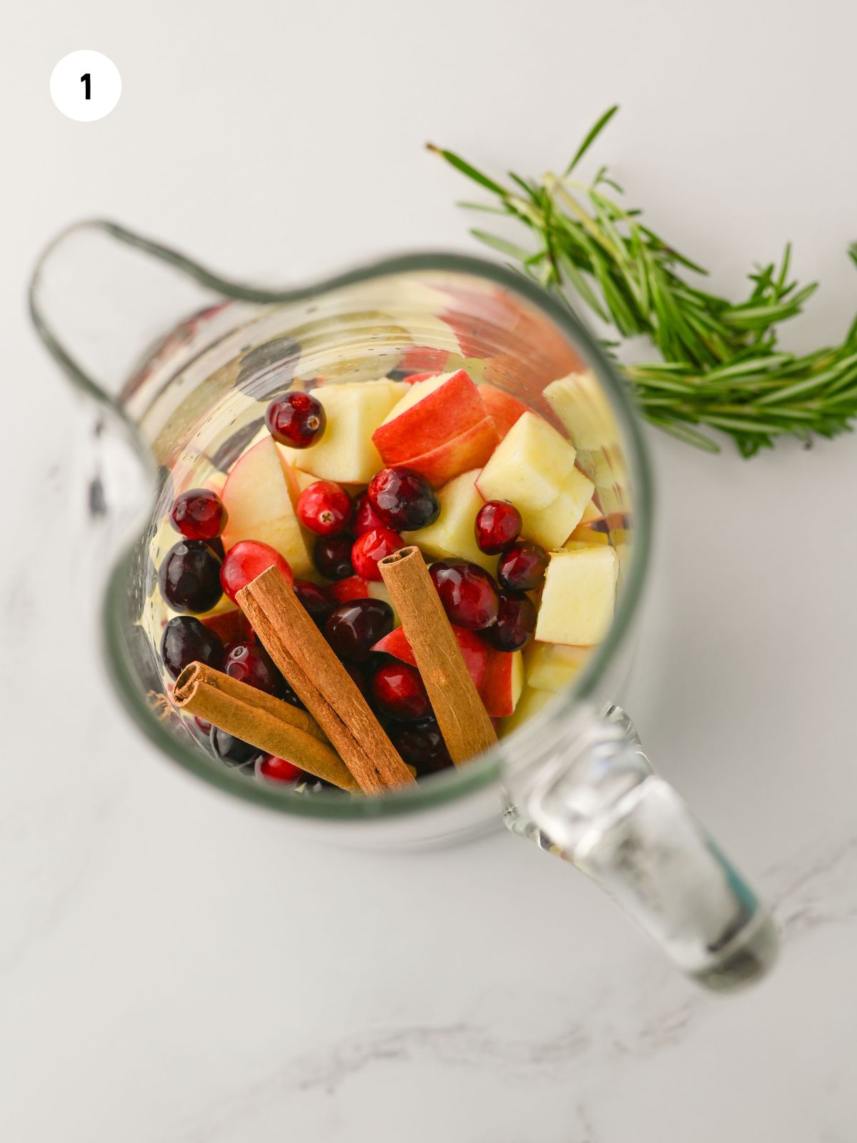 Fresh cranberries, apples, and cinnamon sticks added to a glass pitcher.