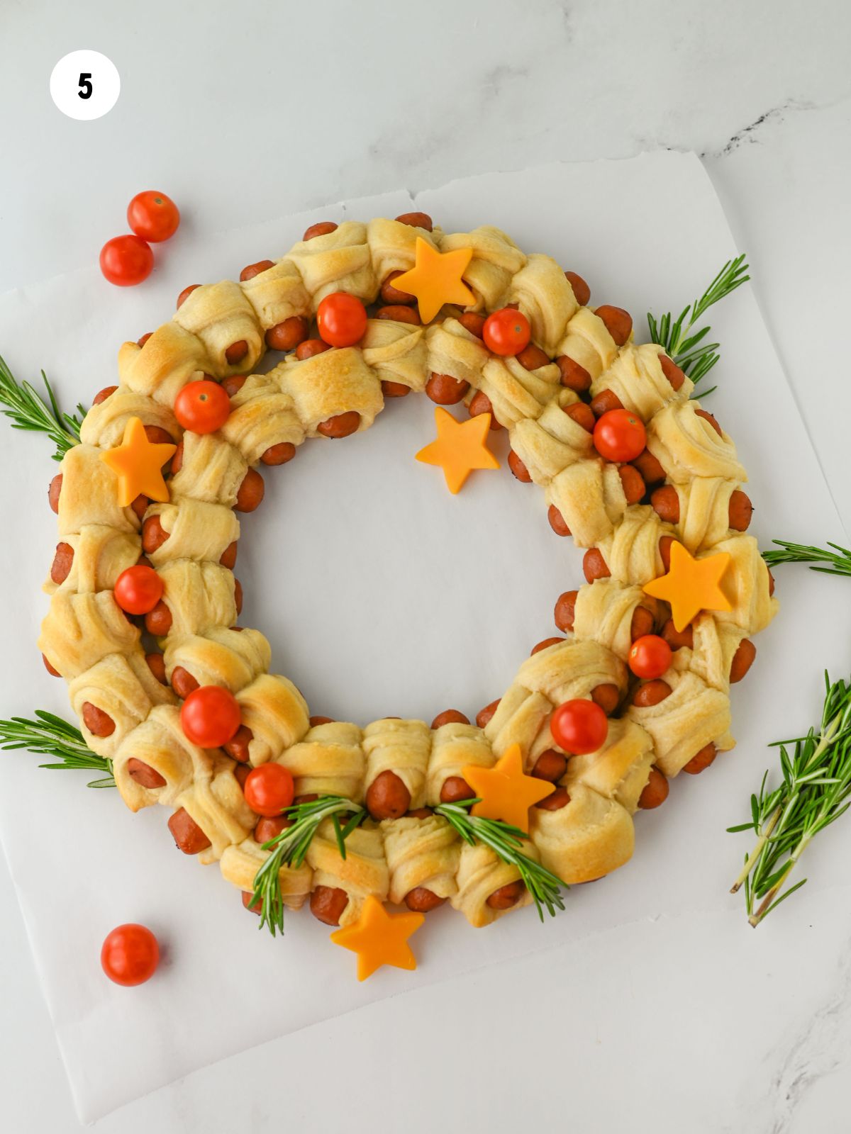 Pigs in a blanket wreath decorated with sprigs of rosemary, cherry tomatoes, and cheddar stars.
