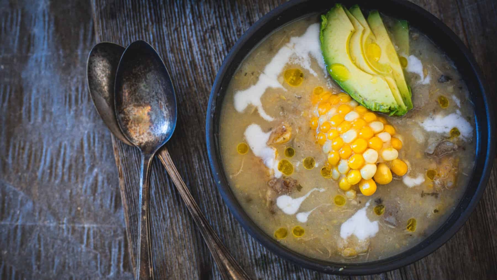 A bowl of ajiaco colombiano soup with chicken, corn on the cob, and avocado.