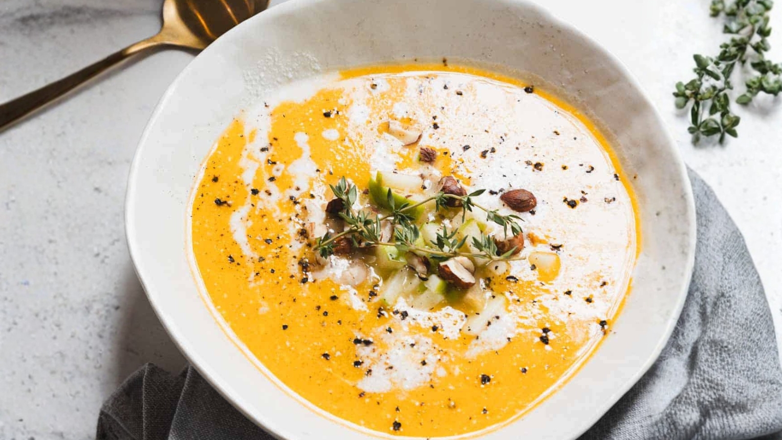 A bowl of creamy orange soup garnished with herbs, chopped nuts, and drizzled cream sits on a marble surface. A small bowl of nuts and sprigs of fresh herbs are nearby, alongside a gray napkin.