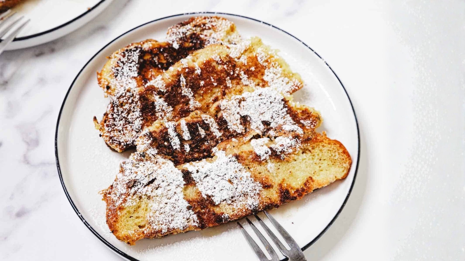 Four slices of French toast topped with powdered sugar on a white plate, with a fork beside them.