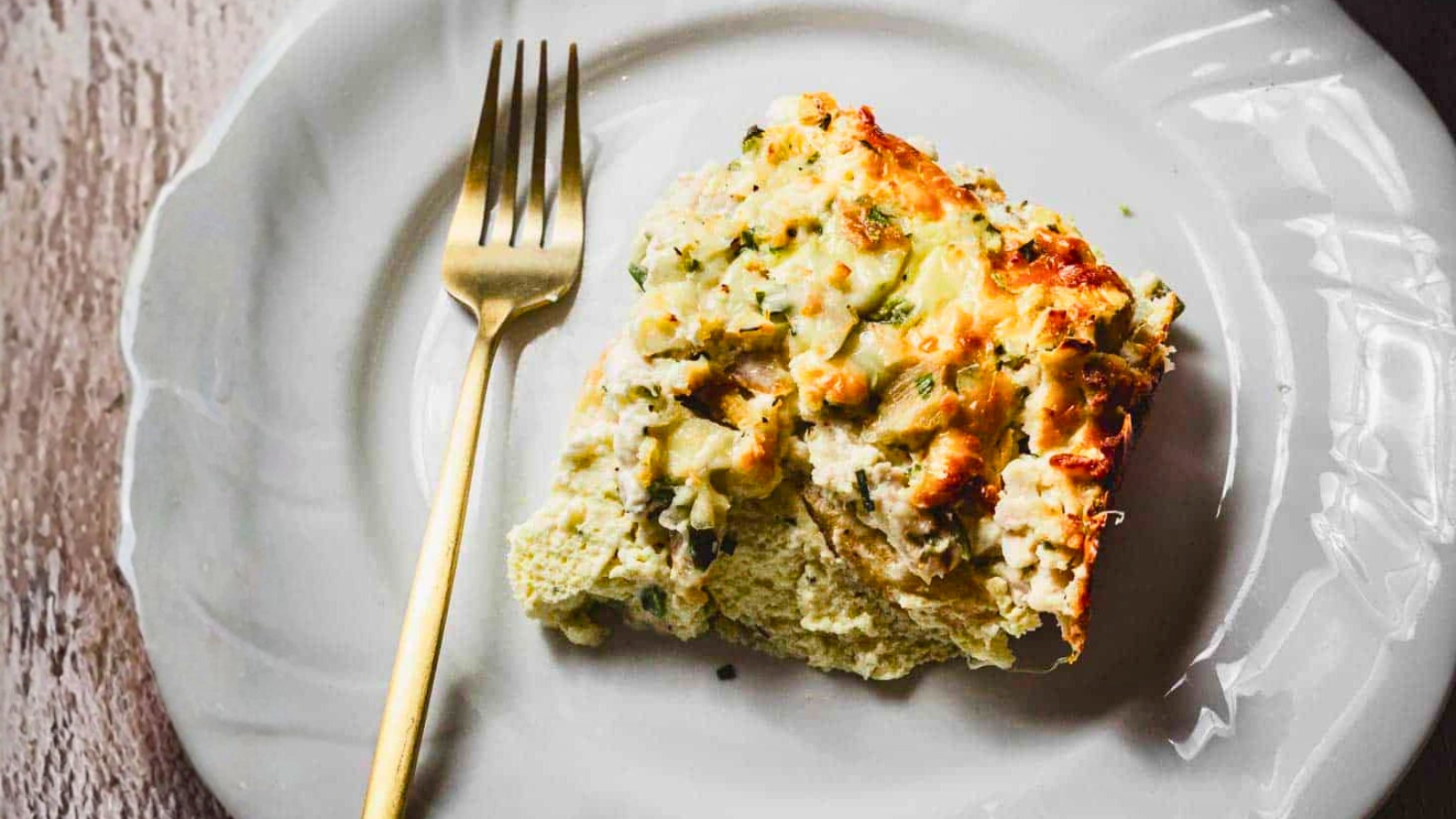 A baked casserole dish filled with a cheesy, golden-brown topping is being scooped with a spatula. The dish appears to contain layers of ingredients beneath the melted cheese, with herbs sprinkled on top for garnish.