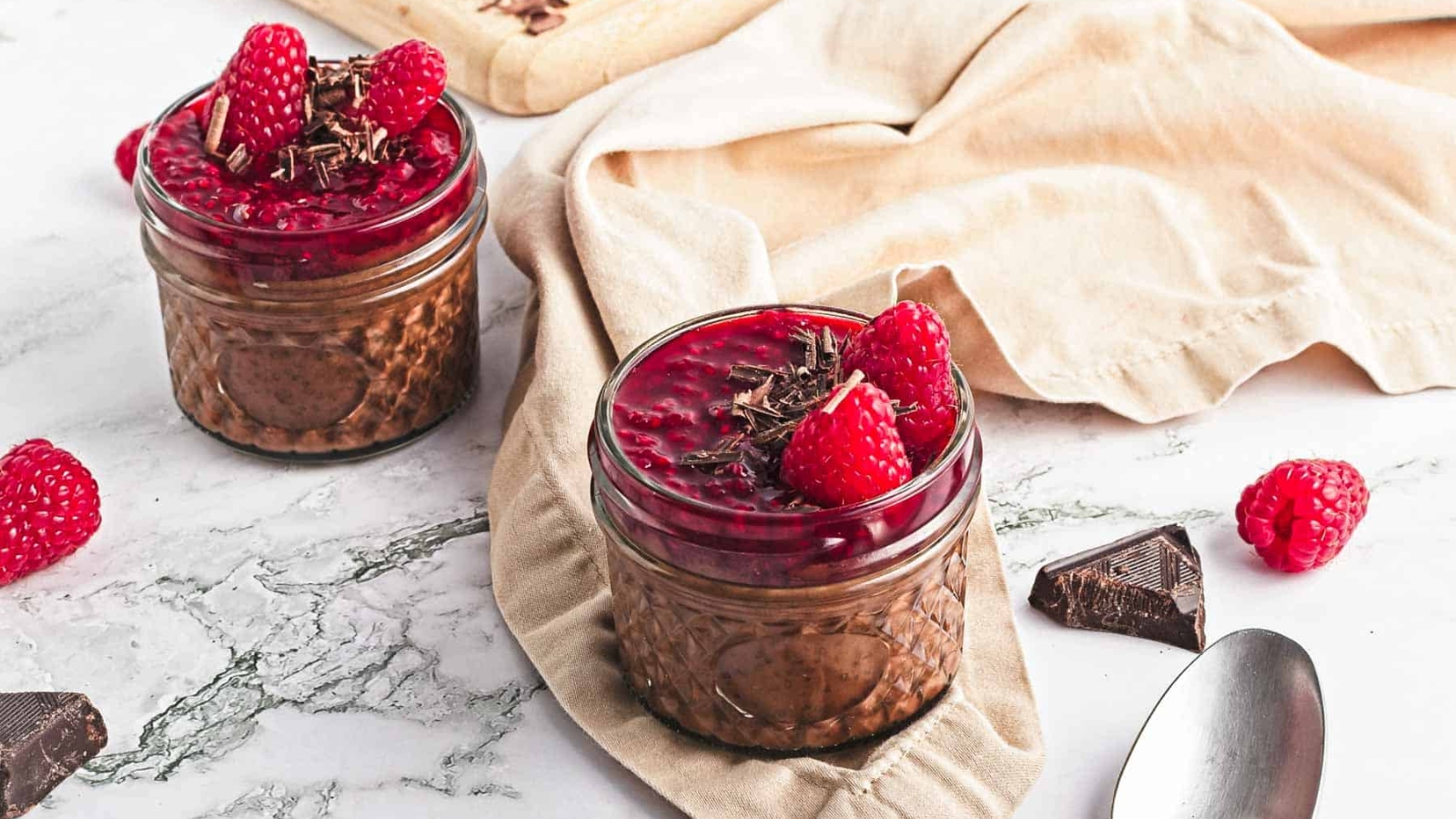 Two small jars filled with chocolate mousse, topped with raspberry sauce, fresh raspberries, and chocolate shavings. A beige cloth, chocolate pieces, loose raspberries, and a spoon are nearby on a marble surface.