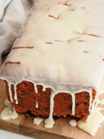 A loaf cake with white icing drips on a wooden board, next to two forks and some dried fruit pieces.
