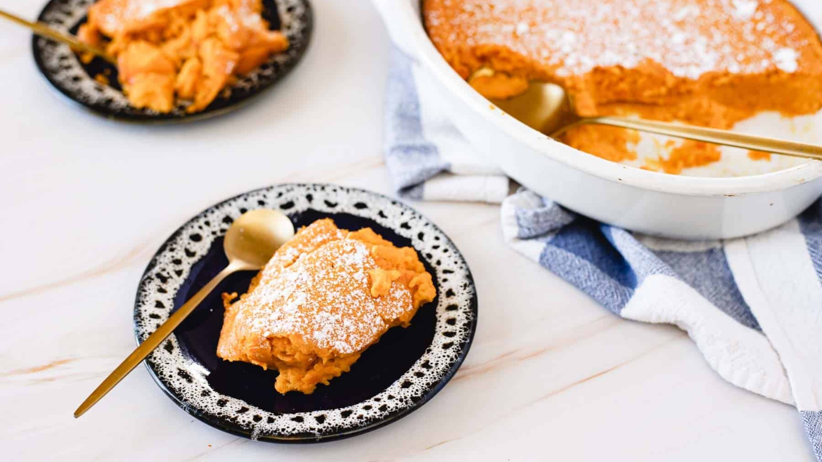 A slice of sweet potato casserole with a dusting of powdered sugar is served on a black and white plate. A gold spoon rests beside it. In the background is a casserole dish with more of the dessert and a white and blue towel.
