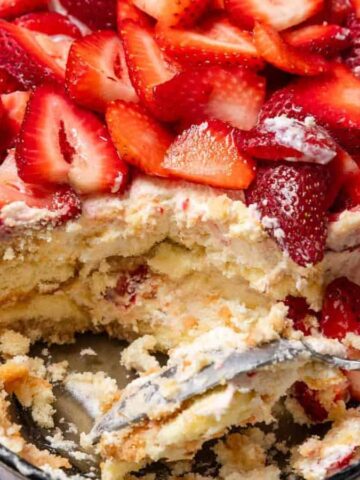 A dish of strawberry tiramisu with a spoon and a portion already served, topped with fresh sliced strawberries.