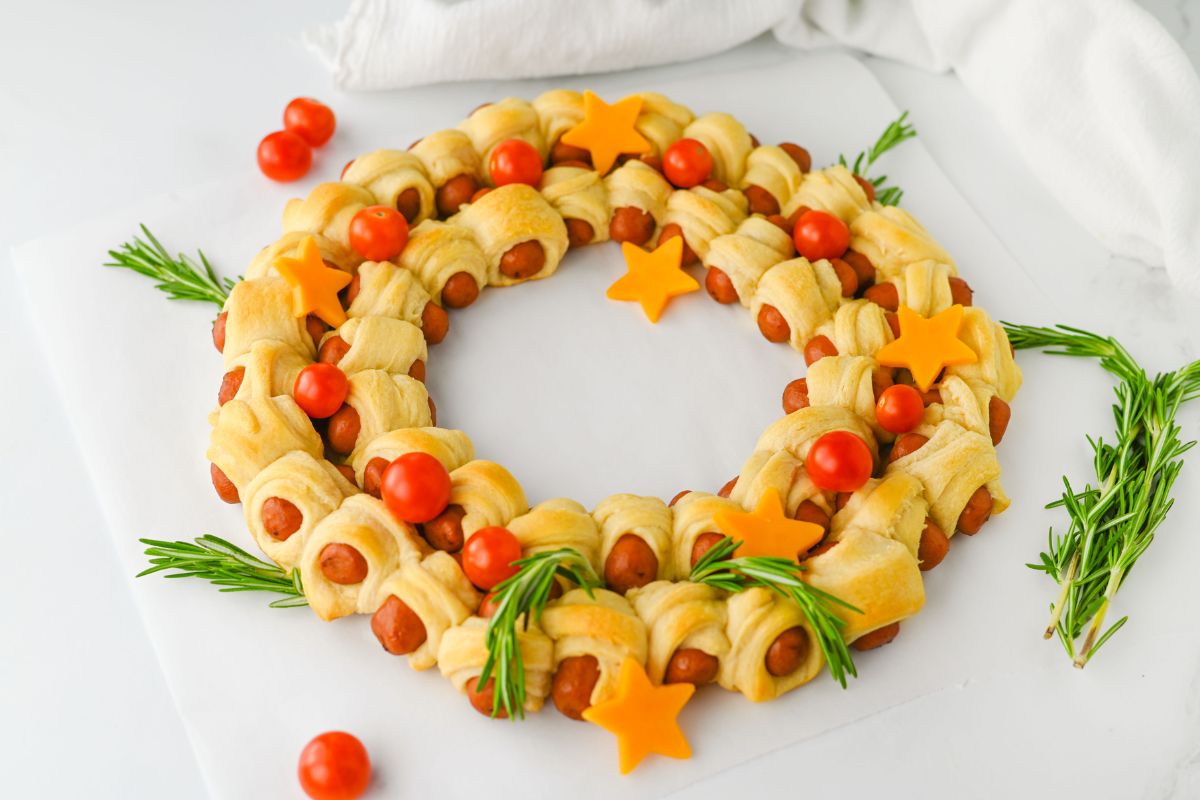 Pigs in a Blanket Christmas Wreath Appetizer decorated with cheese stars, rosemary sprigs, and cherry tomatoes.